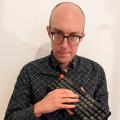 Chris Chinchilla profile image: bald man wearing glasses, dark shirt and holding a keyboard
